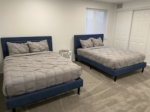 A bedroom with two beds, a white nightstand, a window with blinds, and a white door.