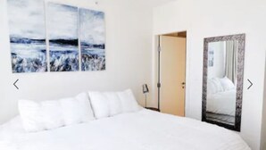 A bedroom with a large bed, white bedding, a nightstand, a lamp, and a door leading to another room. There are three wall-mounted paintings above the bed.