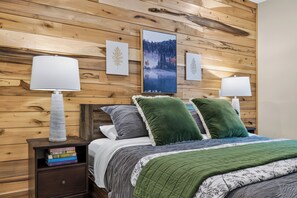 Ein Schlafzimmer mit einer Holzwand, einem Bett mit grün-grauem Bettzeug, zwei Nachttischlampen und einem Bücherregal mit Büchern.