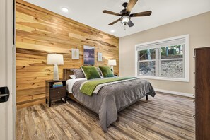 A bedroom with a wooden wall, a bed with a green throw, two bedside lamps, and a ceiling fan. - Sam's Place - Custom 6BR Home - Arcade, Games, Movie Room & Hot Tub! (Massanutten)