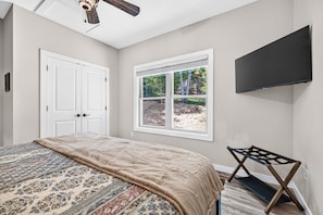 A bedroom with a bed, a window with a view of trees, a ceiling fan, and a mounted television.