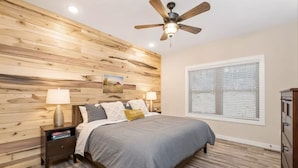 A bedroom with a wooden wall panel, a bed with gray bedding, two bedside lamps, a ceiling fan, a window with blinds, and a nightstand with books.