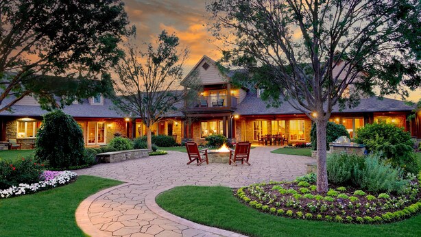 Una casa grande de dos pisos con un patio de piedra, mobiliario exterior y una chimenea.