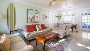A spacious living room with a ceiling fan, a surfboard, and a beach-themed painting.