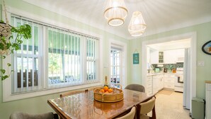 A dining area with a wooden table, chairs, and a fruit basket. There are two hanging lights and a window with blinds.