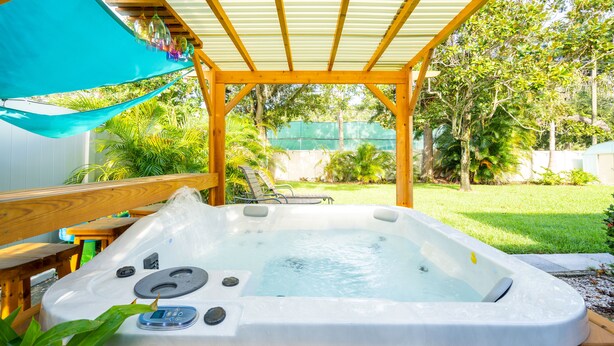 A hot tub under a wooden pergola with a view of a garden and a green fence.