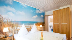 A bedroom with a beach mural, a wooden bed frame, white bedding, a bedside table with a lamp, and a bottle of wine.