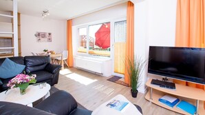 A modern living room with a sofa, coffee table, and a flat-screen TV. There is a dining area with a table and chairs, and a large window with curtains.