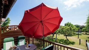 A balcony with a red umbrella, green chairs, and a table with a bottle and a bowl of fruit.