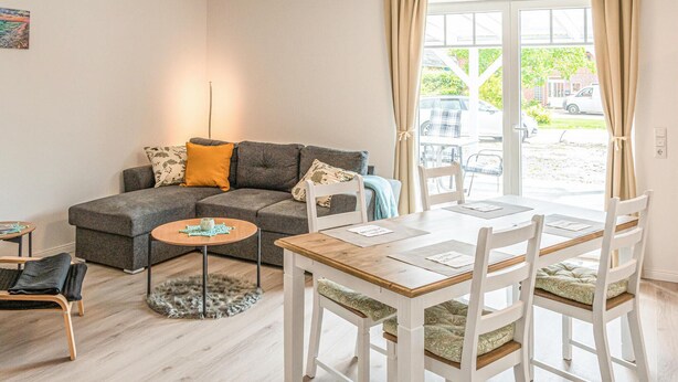 Un salon chaleureux avec un canapé gris, une petite table ronde, une table à manger avec des chaises et une vue sur l’extérieur grâce à de grandes fenêtres.