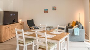 Un salon moderne avec une table à manger en bois, des chaises blanches, un canapé gris et un téléviseur posé sur un meuble en bois.