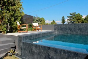 A modern outdoor swimming pool with a stone wall, two wooden chairs, and a wooden deck.