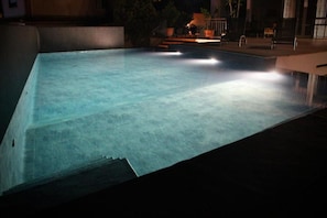 Une piscine extérieure éclairée la nuit, entourée d’un espace sombre et agrémentée de quelques meubles de jardin.