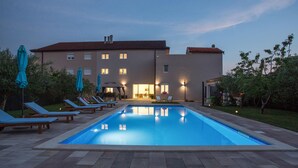 En swimmingpool med liggestole og parasoller, en bygning med mange vinduer og et rødt tag, og et veloplyst udendørs område.