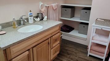 Une salle de bain avec un meuble-lavabo en bois, un lavabo blanc et une fenêtre avec des stores. On trouve des étagères au-dessus du meuble-lavabo et une petite armoire blanche sur la droite.