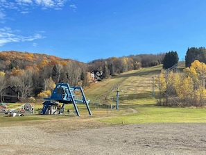 Ein Skigebiet mit einem blauen Sessellift, einem kleinen Gebäude und einem grasbewachsenen Hügel mit Bäumen im Hintergrund.