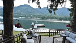 A deck with outdoor seating overlooking a lake, a dock with a boat, and mountains in the background.
