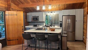 A compact kitchen with a central island, bar stools, a microwave, a refrigerator, and a window overlooking greenery.