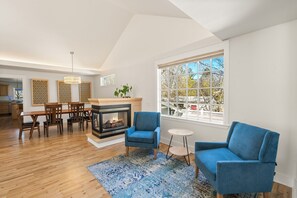 A spacious living room with a fireplace, blue armchairs, a dining area with wooden furniture, and a large window offering a view of trees. - The Ashland Social | Main House (Ashland)