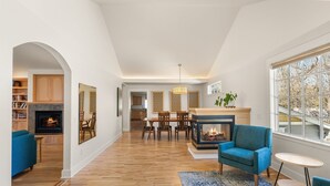 Een ruime woonkamer met een open haard, een eethoek en uitzicht door grote ramen.
