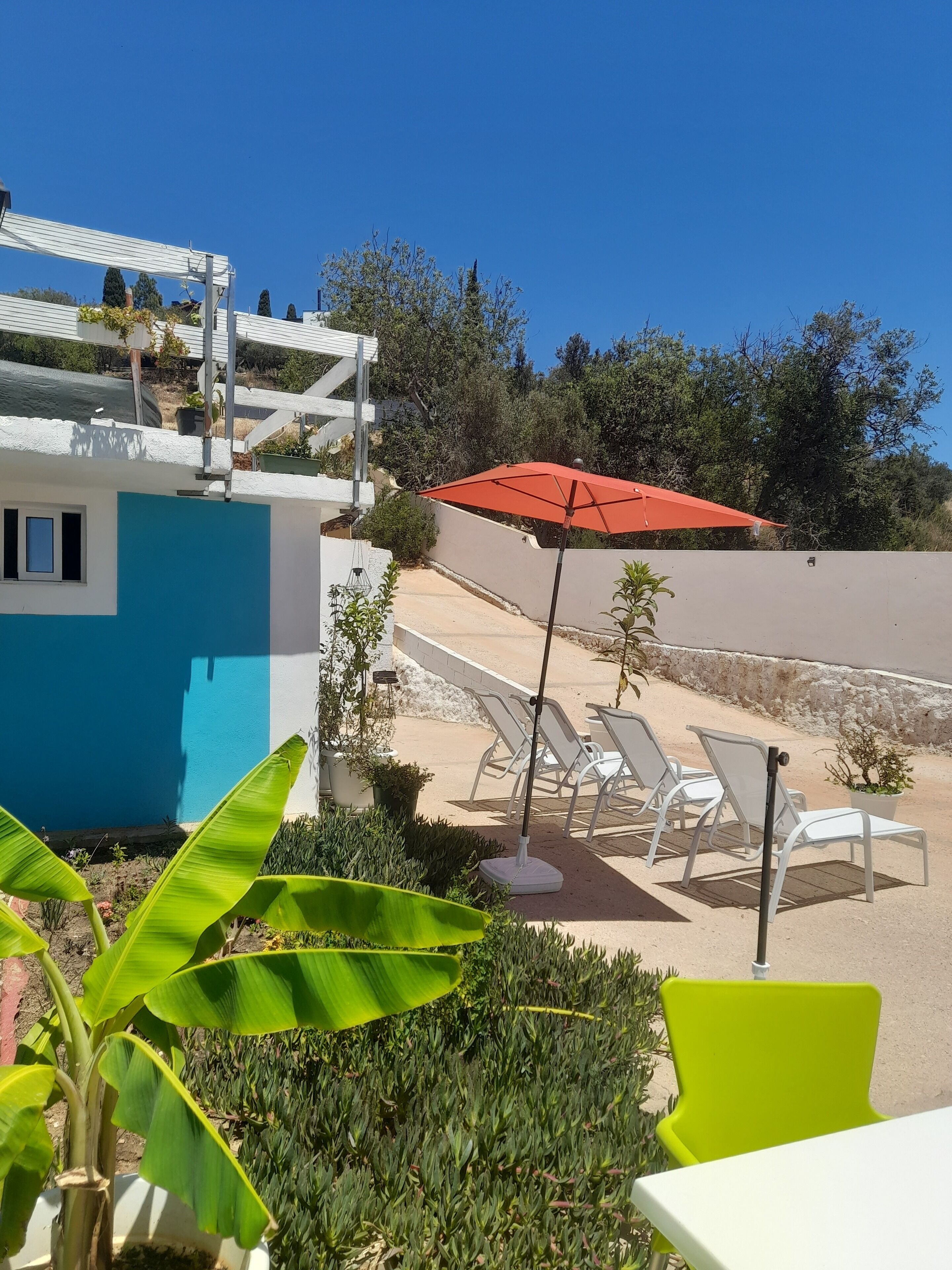 A patio area with white lounge chairs and a red umbrella, surrounded by greenery and a white building with blue accents.