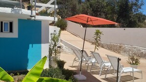 A patio area with white lounge chairs and a red umbrella, surrounded by greenery and a white building with blue accents. - Studio in Gale Albufeira (Albufeira)