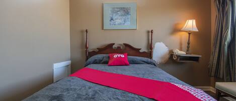 Een net opgemaakt bed met een rode plaid, een houten hoofdbord en een nachtkastje met een lamp en telefoon.