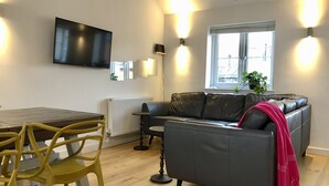 Un salon moderne avec une table en bois, des chaises jaunes, un canapé gris agrémenté d’un plaid rose et un téléviseur fixé au mur.