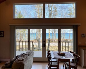 A room with a large sliding glass door leading to a deck with a view of the sea and trees. Inside, there is a wooden table with chairs and a sofa. - 2-bed cottage on Cable's Bay with private beach (Beaver Island)