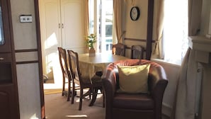 Un coin repas avec une table ronde, des chaises et un canapé. Il y a une horloge au mur et une fenêtre avec des rideaux.