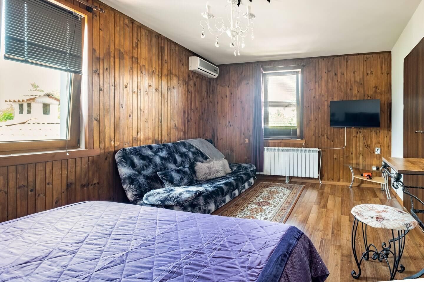 Een kamer met houten wanden, een bed met een paars sprei, een bank met een zachte hoes, een aan de muur bevestigde televisie en een radiator.