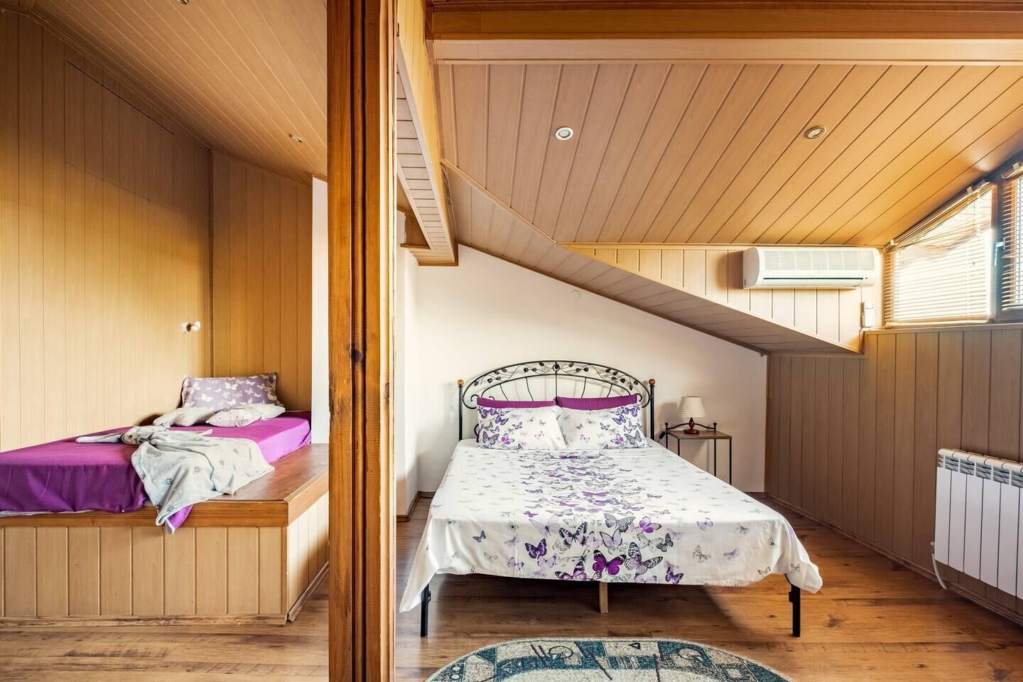 A bedroom with a sloped ceiling, wooden walls, a bed with a floral bedspread, a bench with a purple cover, and a radiator.