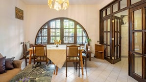 A dining area with a round table, chairs, a chandelier, and large windows.