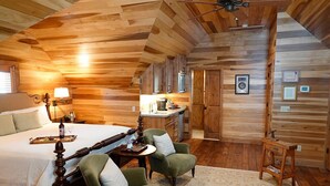 Une chambre à coucher dans une cabane en bois, avec un lit à baldaquin, une petite table avec un plateau, deux fauteuils verts, une table de chevet en bois et un ventilateur de plafond.