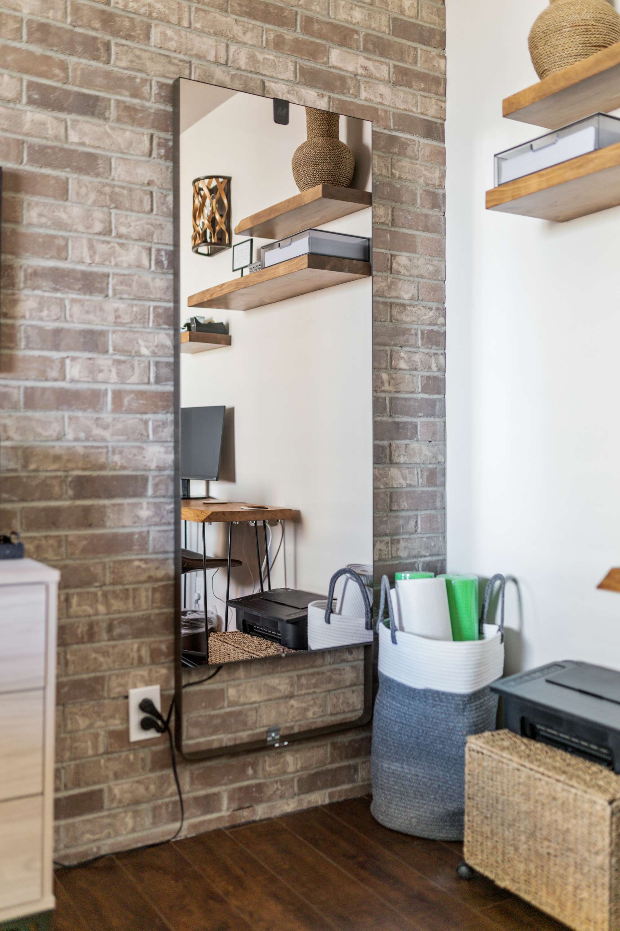 A room with a brick wall, a large mirror, a desk with a computer, shelves with decorative items, and a basket with office supplies.