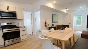 Une cuisine moderne équipée d’appareils électroménagers en acier inoxydable, un coin repas avec une table en bois et des chaises blanches, et un salon avec un canapé et un téléviseur.