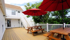 Une terrasse en bois avec des tables de pique-nique et des parasols rouges, des rampes blanches et un escalier menant à un deuxième niveau.