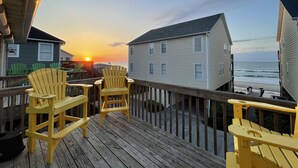 Una terraza de madera con sillas Adirondack amarillas que dan a una playa con casas al fondo.