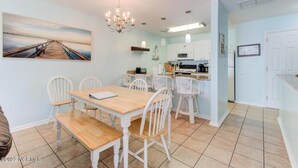 Un comedor con mesa de madera y sillas blancas, una cocina con armarios blancos, y un cuadro de un muelle en la pared.