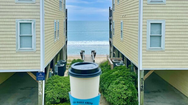 Vista de una propiedad frente a la playa con dos casas, una terraza de madera y una taza de café en primer plano.