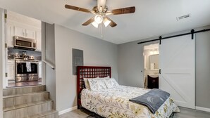 A bedroom with a bed, a ceiling fan, a wooden headboard, and a view of a kitchen and bathroom through an open door.