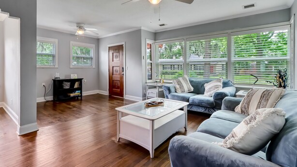 A spacious living room with hardwood floors, a ceiling fan, and large windows offering a view of greenery outside.