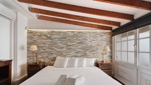 Una cama bien hecha con ropa de cama y toallas blancas, pared con textura de piedra, vigas de madera y un ventanal con persianas.