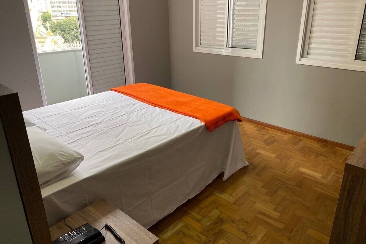 A bedroom with a bed, an orange throw blanket, wooden flooring, and white blinds.
