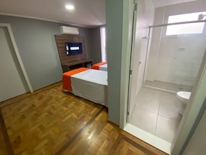 Una habitación con piso de madera, una cama con colcha naranja, un televisor en una pared de madera y un área de ducha con inodoro.