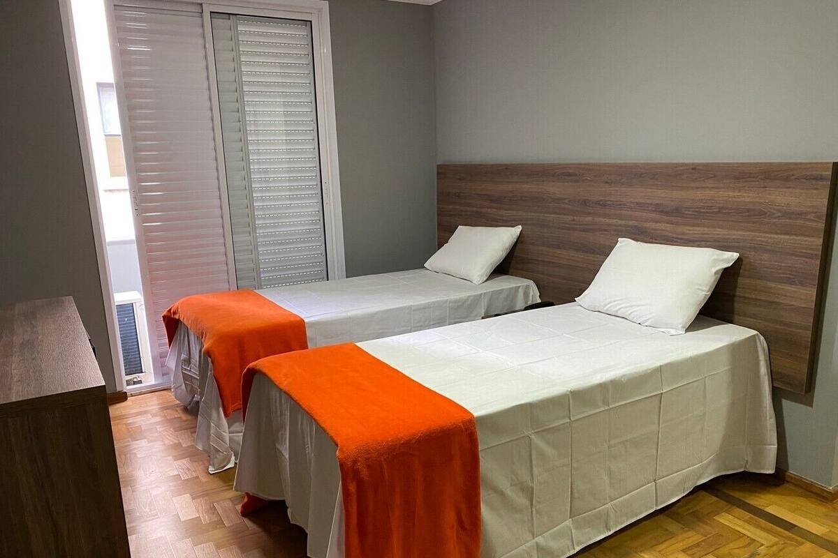 A hotel room with two beds, each with a white bedspread and an orange runner. The room has wooden flooring and a grey wall with a wooden headboard.