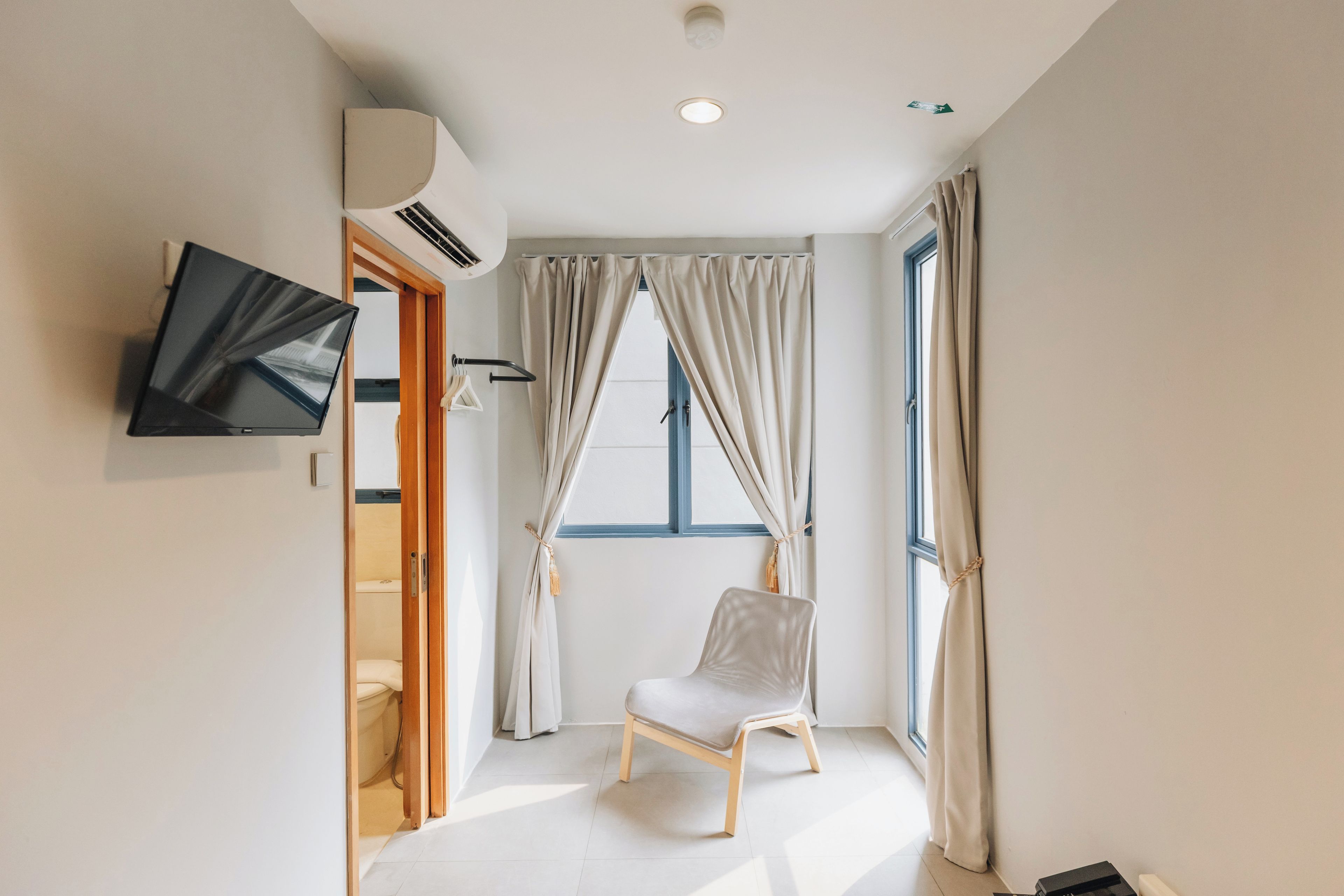Una habitación moderna con televisor de pantalla plana en la pared, una ventana con cortinas, una silla blanca y una puerta de madera.