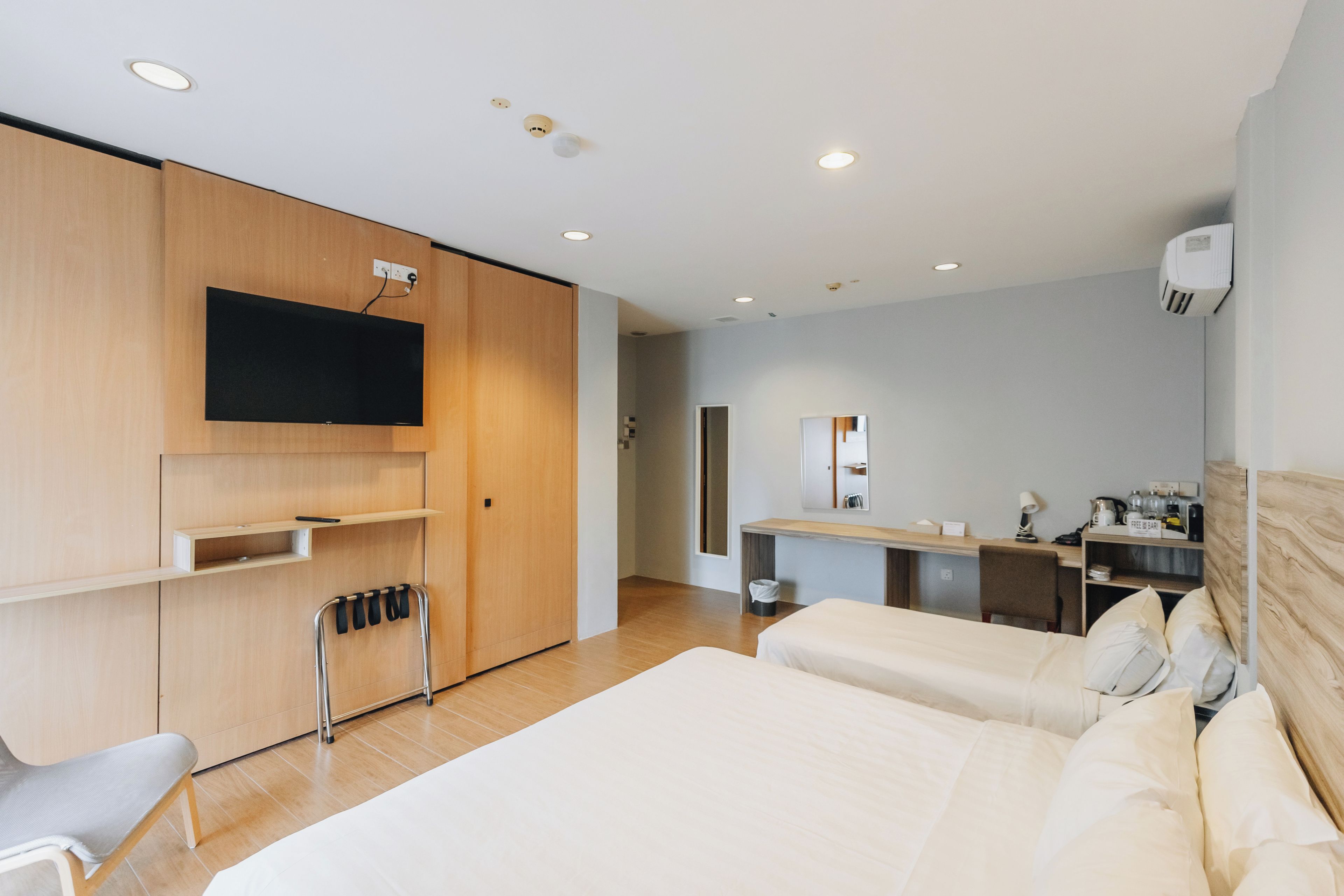 A modern hotel room with a large bed, a TV mounted on a wooden panel, a desk with a mirror, and a small kitchenette.