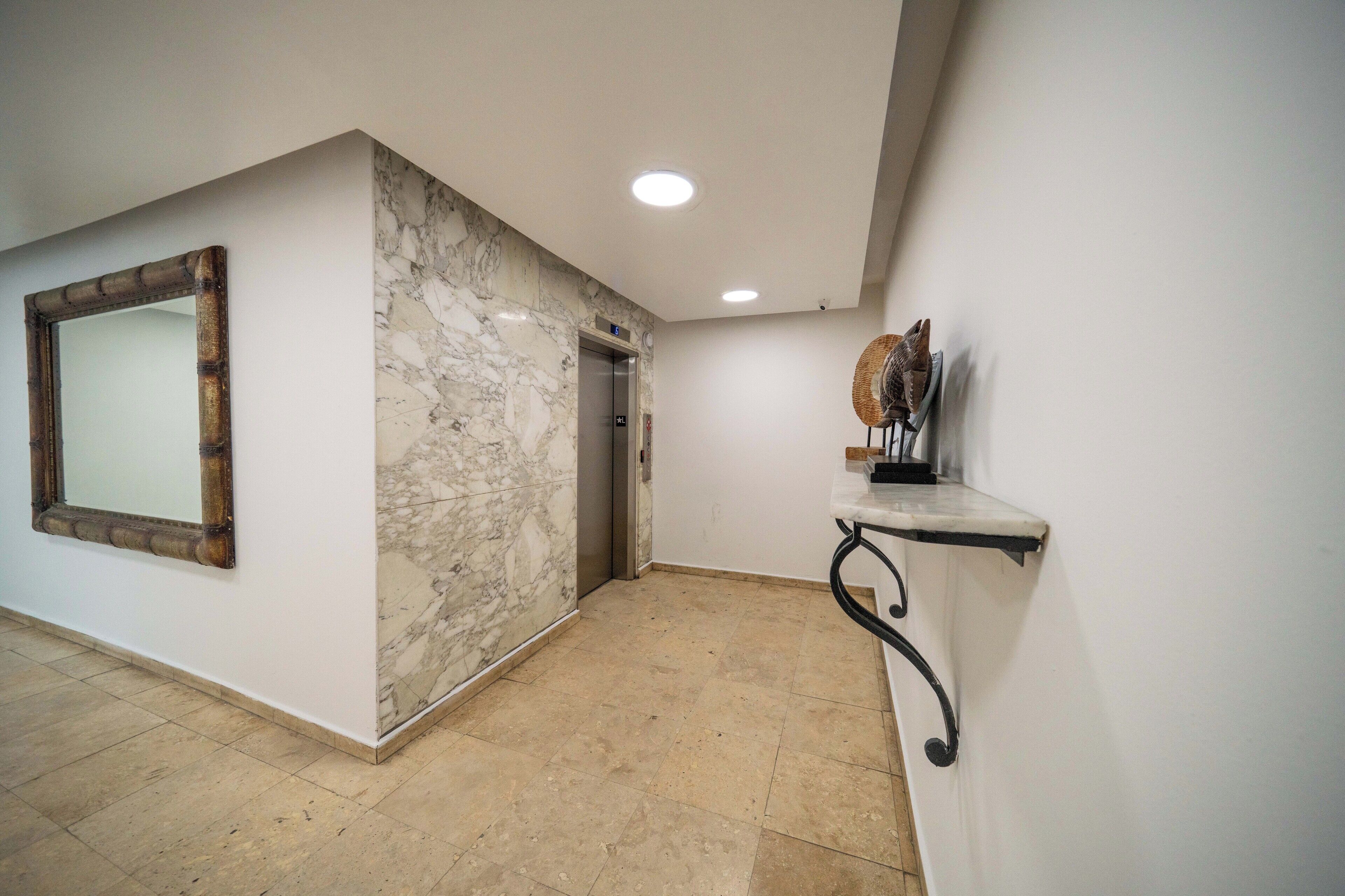 Un couloir avec un mur de marbre, un miroir, un ascenseur et une étagère sur laquelle se trouve un ventilateur.
