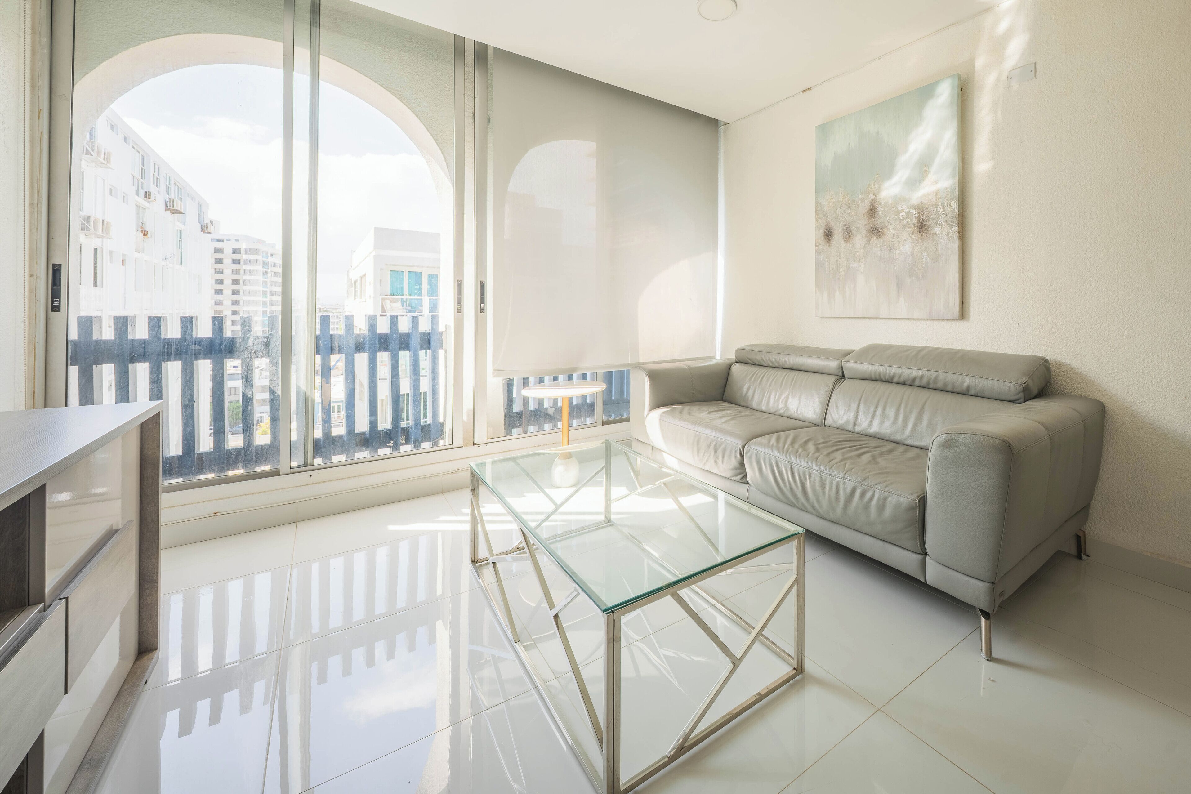 A modern living room with a glass coffee table, a beige sofa, and a large window offering a view of buildings.